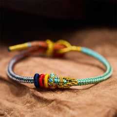 Five-Color Eight Thread Peace Knot Bracelet – Gradient turquoise gray multicolor woven cord with gold yellow red blue decorative knots, tan fabric low side angle