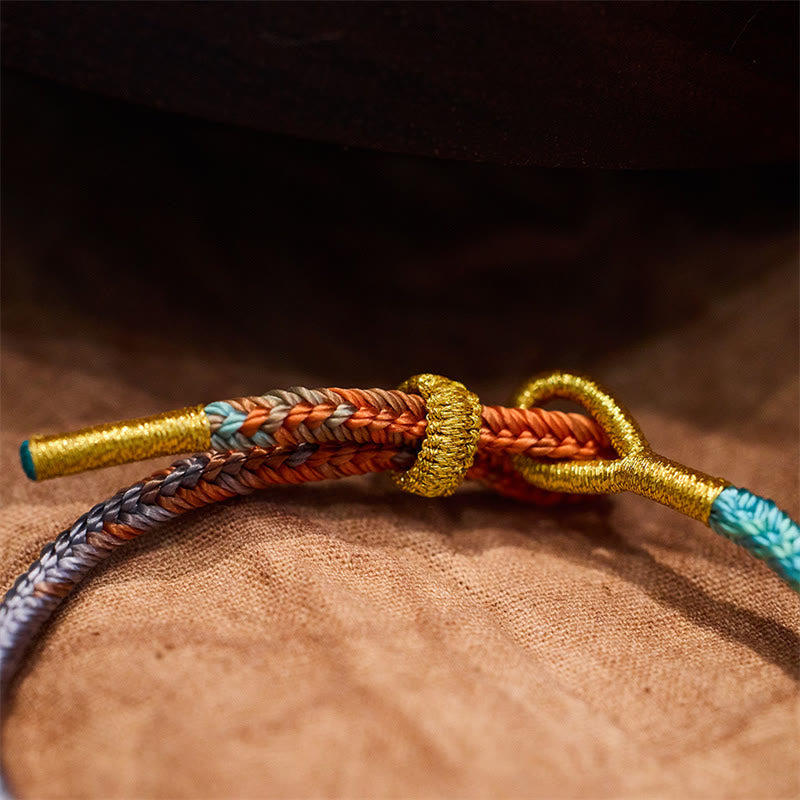 Five-Color Eight Thread Peace Knot Bracelet – Orange brown gray sky blue braided cord with gold metallic thread wraps and sliding knot closure, tan fabric low angle