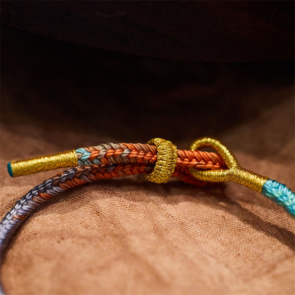 Five-Color Eight Thread Peace Knot Bracelet – Orange brown gray sky blue braided cord with gold metallic thread wraps and sliding knot closure, tan fabric low angle