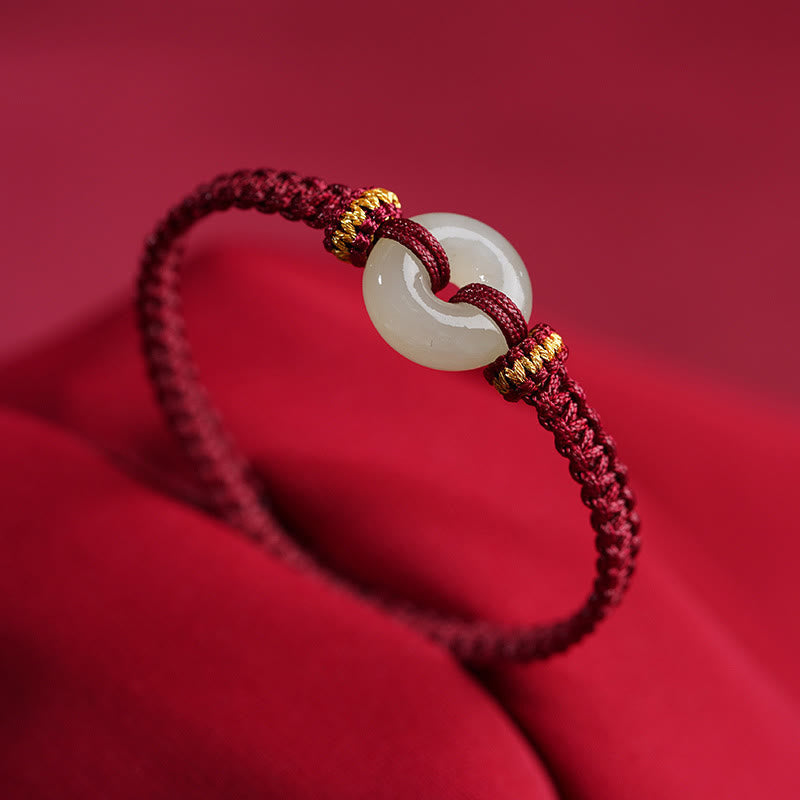 Peace Buckle Jade Abundance Bracelet – Burgundy braided cord with pale celadon jade peace buckle and gold-wrapped knots, crimson fabric background three-quarter