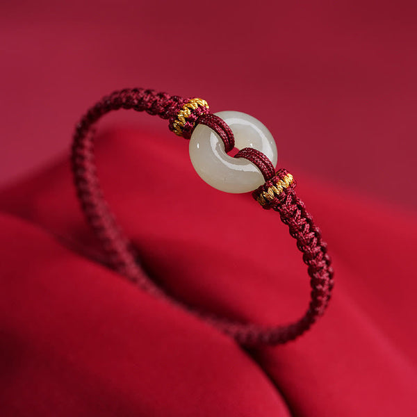 Peace Buckle Jade Abundance Bracelet – Burgundy braided cord with pale celadon jade peace buckle and gold-wrapped knots, crimson fabric background three-quarter