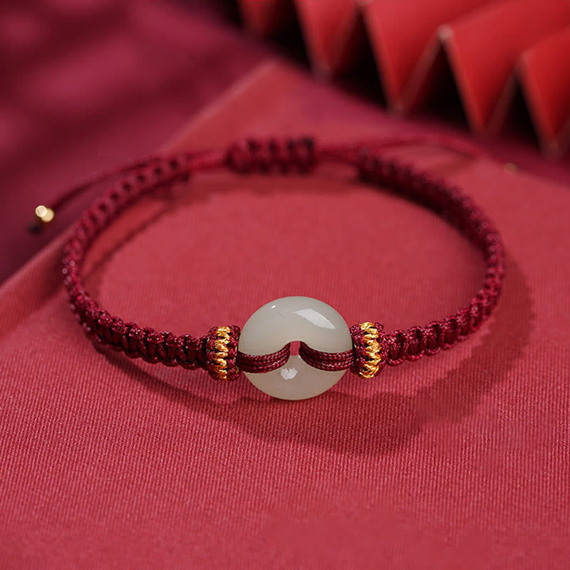 Peace Buckle Jade Abundance Bracelet – Deep red braided cord with pale green jade donut bead and gold metal rings, soft red fabric with geometric bokeh, elevated side
