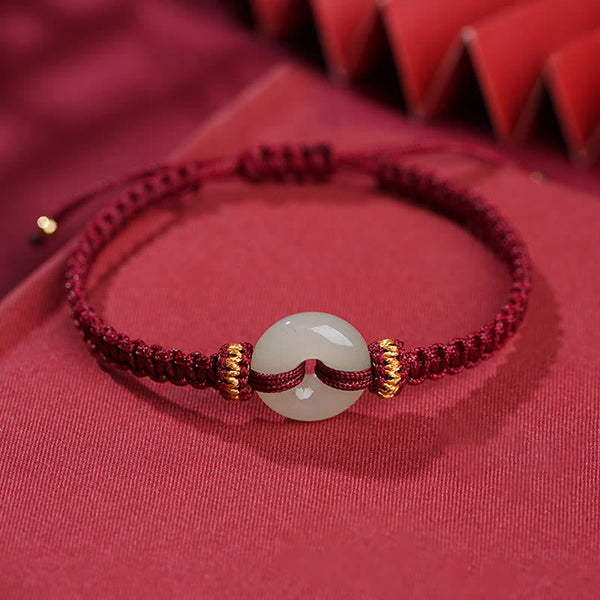 Peace Buckle Jade Abundance Bracelet – Deep red braided cord with pale green jade donut bead and gold metal rings, soft red fabric with geometric bokeh, elevated side