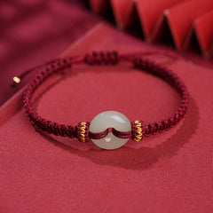 Peace Buckle Jade Abundance Bracelet – Deep red braided cord with pale green jade donut bead and gold metal rings, soft red fabric with geometric bokeh, elevated side