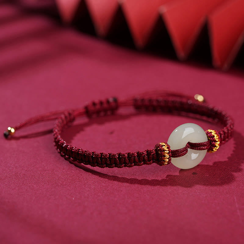 Peace Buckle Jade Abundance Bracelet – Burgundy red cord with white jade peace buckle and gold accents on matching red fabric, low side angle shallow depth