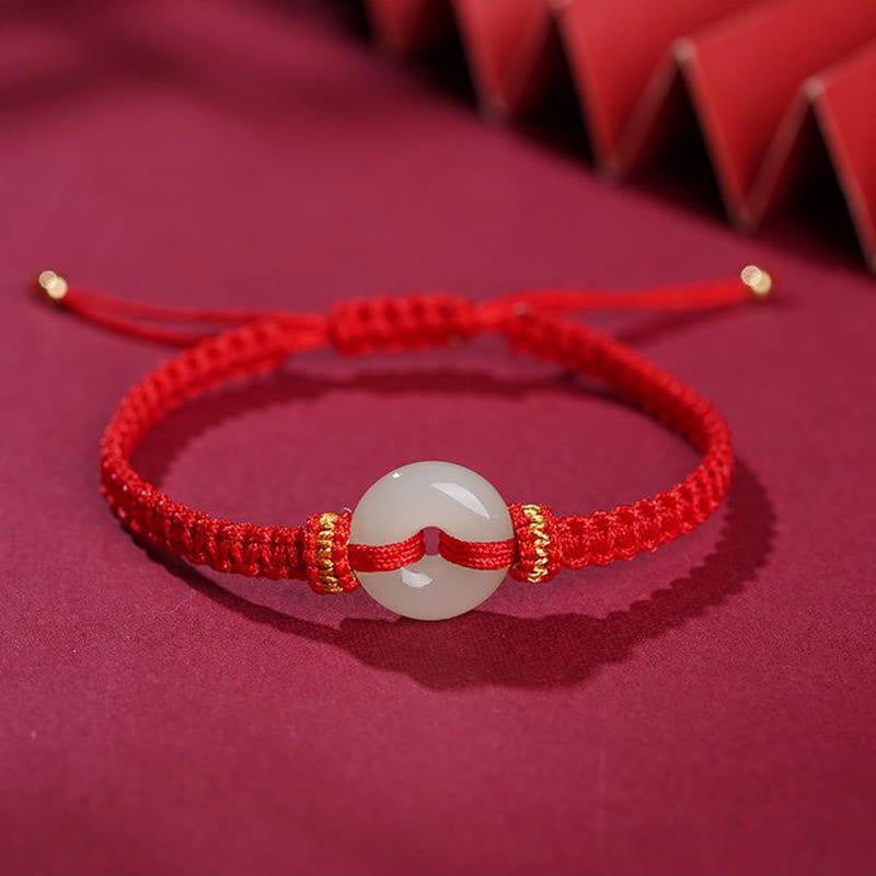 Peace Buckle Jade Abundance Bracelet – Red braided cord with white jade disc and gold-toned accents on deep red fabric with folded fan decoration, elevated angle