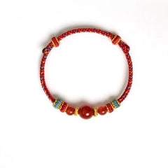 Dunhuang Agate Bracelet – red braided cord bracelet with large central carnelian bead and gold accents on white background, grounding jewelry