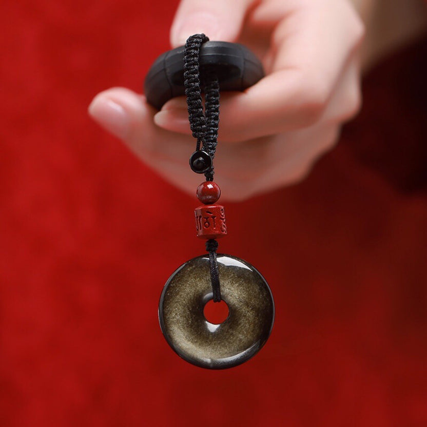 Gold Obsidian Pendant – Hand holding gold sheen obsidian keychain with red bead against red background, protection charm Imagen principal del producto