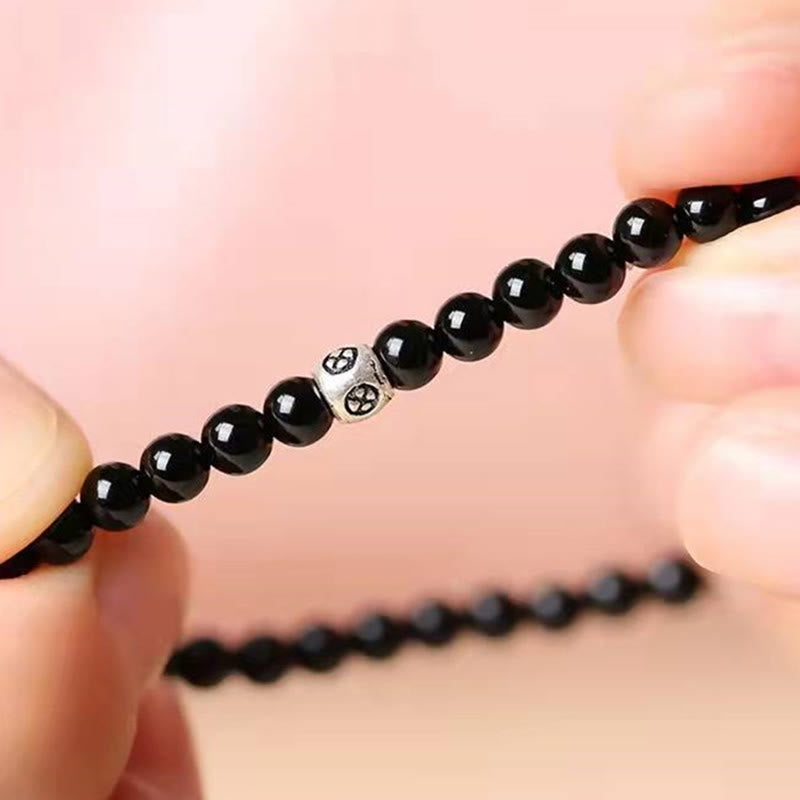 Black Obsidian Smoky Quartz Bracelet – Two hands holding black beaded bracelet strand taut showing silver decorative accent bead, macro close-up with soft pink bokeh