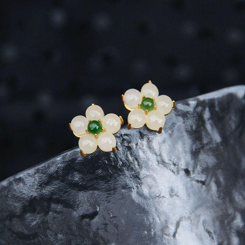 White Jade Flower Blessing Stud Earrings – White jade petal flower studs with green centers in gold metal on gray-white marble surface, dark navy bokeh background