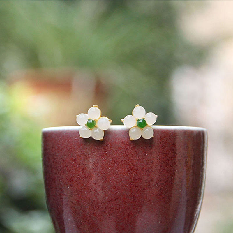 White Jade Flower Blessing Stud Earrings – Cream jade five-petal flower earrings with green centers on gold posts, burgundy ceramic cup with blurred green outdoor background