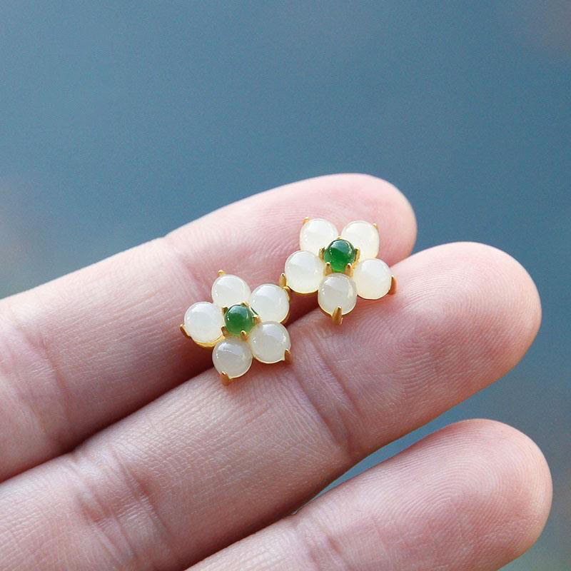 White Jade Flower Blessing Stud Earrings – White jade flower studs with green jade centers in gold settings held on fingertips, blue-gray gradient background, diagonal angle