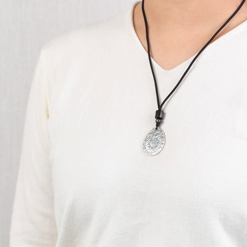 Yin Yang Bagua Water Drop Necklace – Silver teardrop pendant with embossed circular pattern and spiral design on black cord worn with white V-neck, gray background