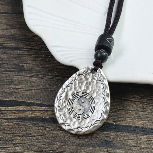 Yin Yang Bagua Water Drop Necklace – Silver teardrop pendant with central yin-yang surrounded by eight trigrams and wave border on dark cord, wood grain surface