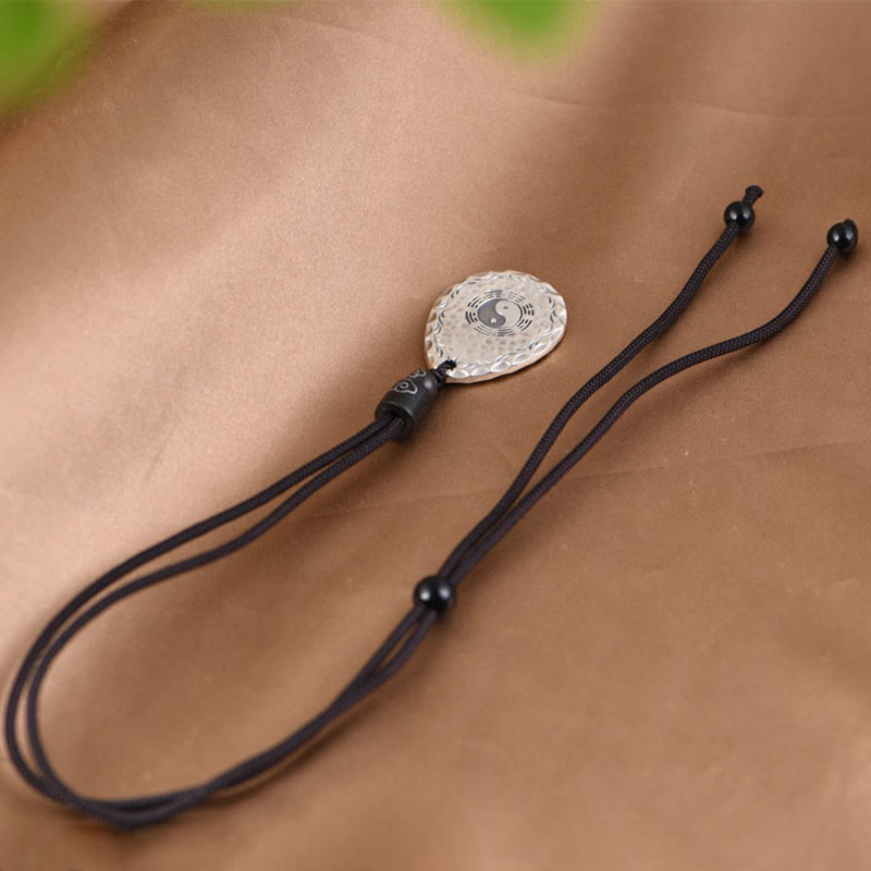 Yin Yang Bagua Water Drop Necklace – Silver circular yin-yang and bagua pendant on black cord with decorative beads on brown fabric with green foliage blur, diagonal