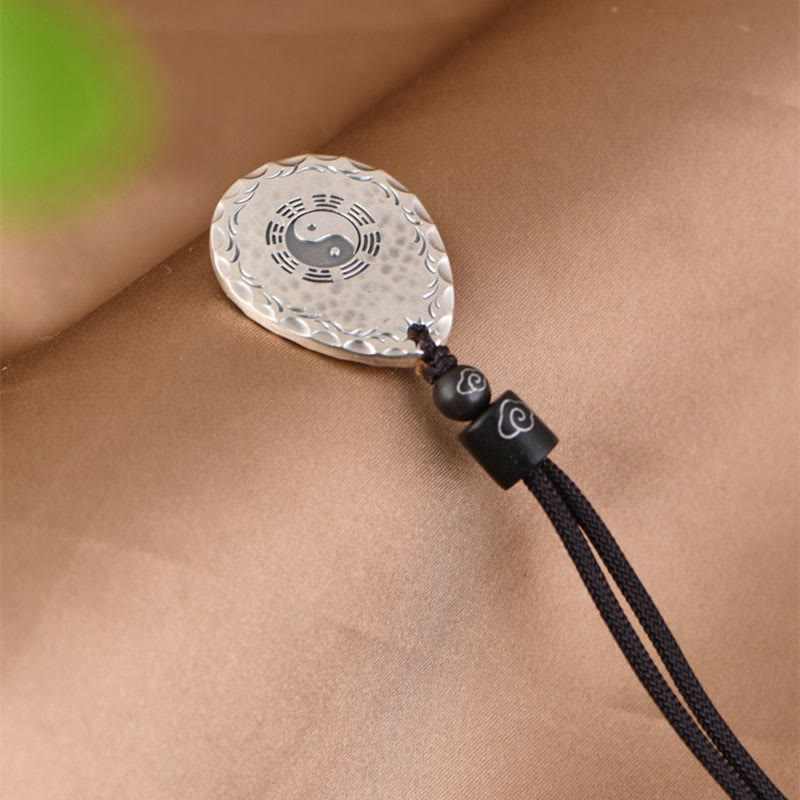 Yin Yang Bagua Water Drop Necklace – Silver teardrop pendant with yin-yang center and bagua trigrams, wave border and cloud-accent beads, tan fabric green blur overhead