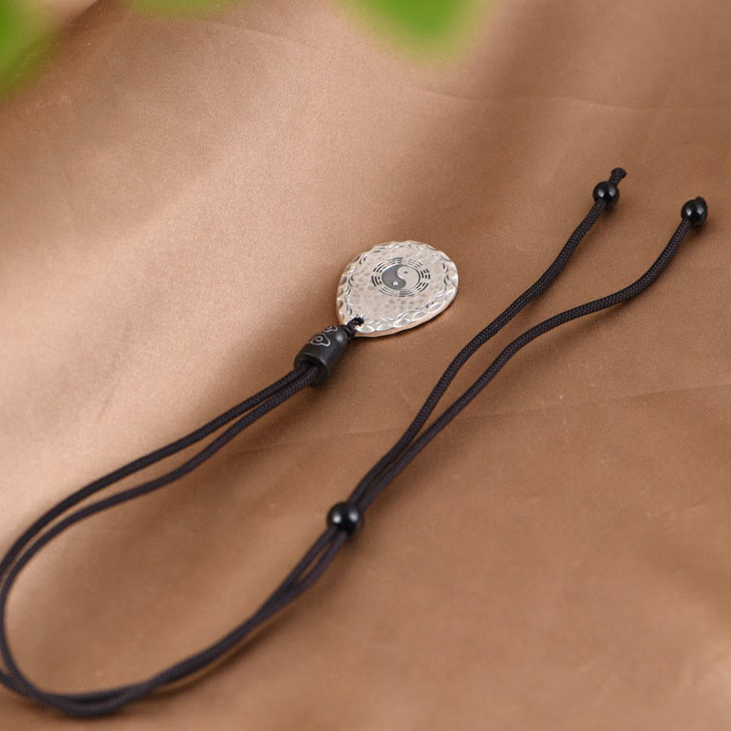 Yin Yang Bagua Water Drop Necklace – Silver circular yin-yang bagua pendant on black cord with wooden beads, brown fabric with green leaves, elevated diagonal