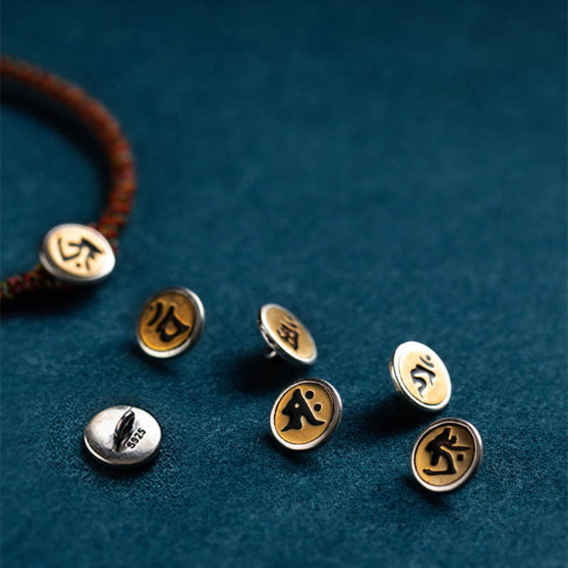 Chinese Zodiac Natal Buddha Bracelet – Six silver-rimmed gold charms with black Sanskrit symbols, one on red cord bracelet, diagonal overhead view