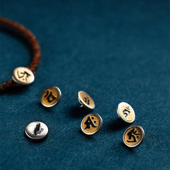 Chinese Zodiac Natal Buddha Bracelet – Six silver-rimmed gold charms with black Sanskrit symbols, one on red cord bracelet, diagonal overhead view