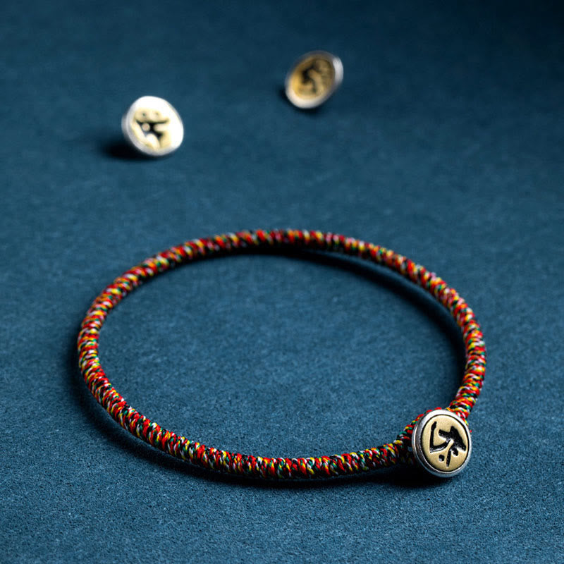 Chinese Zodiac Natal Buddha Bracelet – Red green yellow black cord bracelet with gold symbol bead, shallow depth of field on teal fabric
